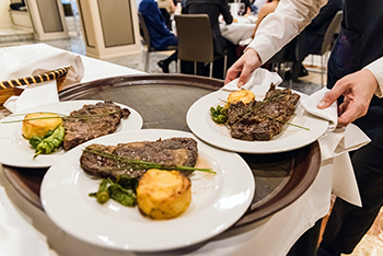 Waiter picks up a plate with a meat steak during a dinner at a restaurant, with unfocused diners in the background.