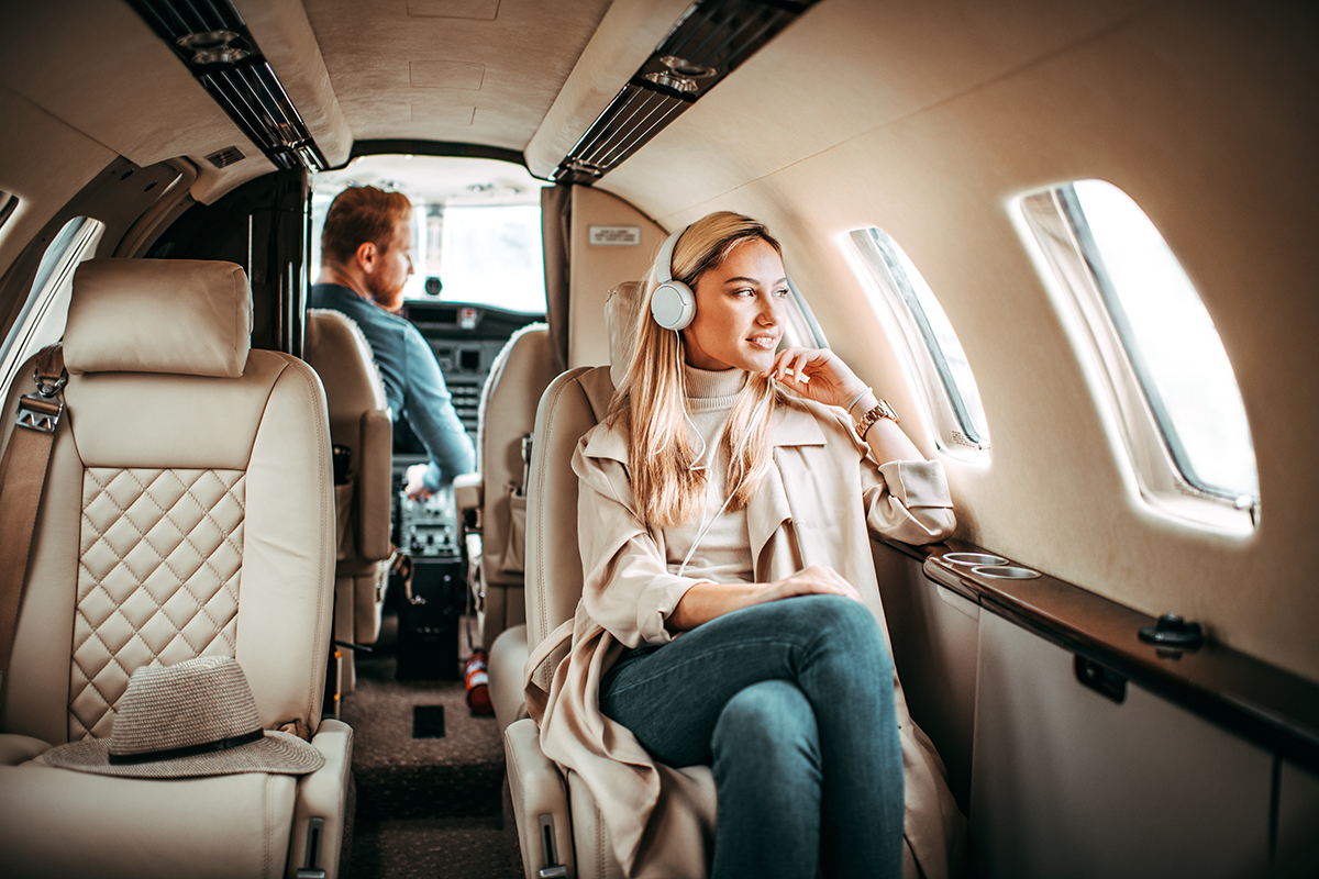 Young successful woman sitting on a private jet and listening to music through headphones. A pilot is sitting in a cockpit.